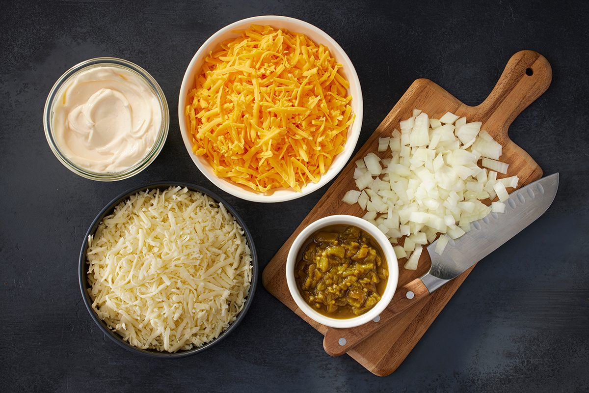 Bowls of shredded cheddar and Monterey Jack cheese, mayonnaise, and chopped pickled jalapeños sit next to a cutting board with diced onions and a knife on a dark surface.