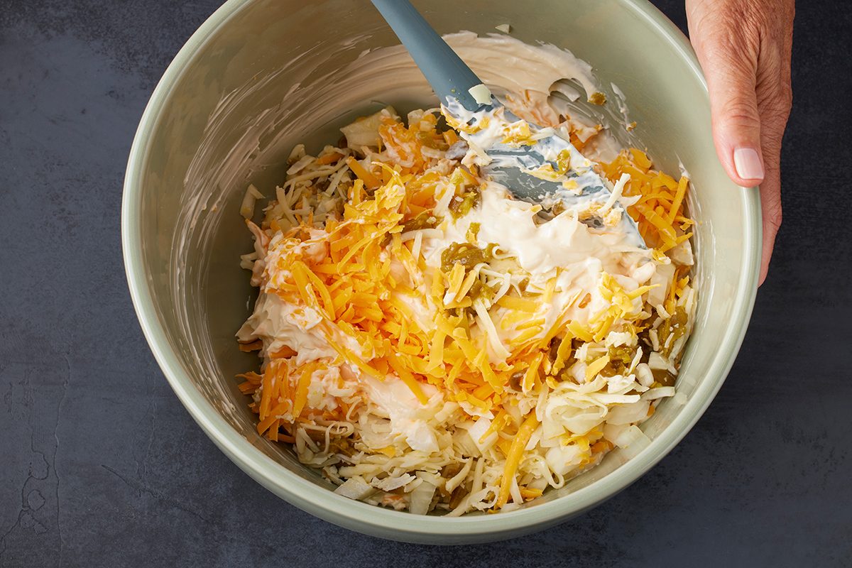 A hand holds a mixing bowl filled with shredded cheese, hash browns, jalapeños, and creamy sauce, being mixed with a spatula on a dark countertop.