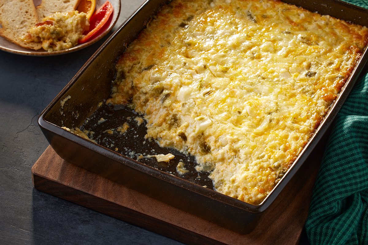 A rectangular baking dish filled with a golden, cheesy casserole sits on a wooden board. Part of the casserole has been served, revealing its creamy interior. A plate with a small portion and sliced tomatoes is nearby.