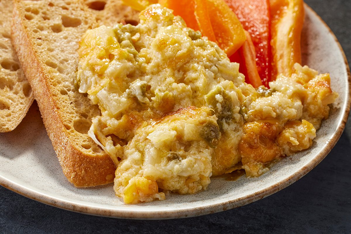 Plate with a serving of cheesy, baked casserole, slices of bread, and orange bell pepper strips. The food looks creamy and slightly golden, garnished with green herbs.