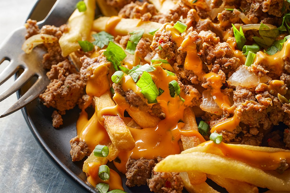 A close-up of loaded fries topped with seasoned ground beef, melted cheese sauce, and chopped green onions and cilantro on a dark plate with a fork on the side.