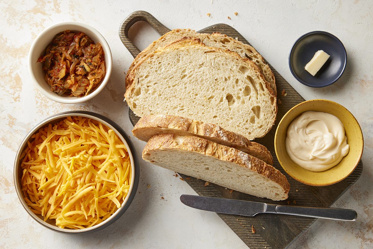 Sliced bread on a cutting board with a knife, next to bowls of shredded cheese, caramelized onions, mayonnaise, and a pat of butter on a small plate.