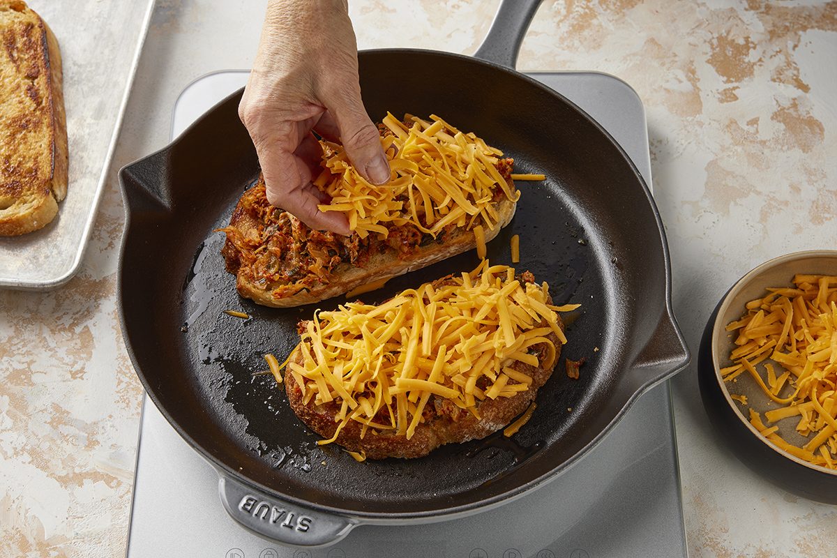 A hand sprinkles shredded cheddar cheese onto two slices of bread with meat in a black skillet, preparing a sandwich. A bowl of cheese and another slice of bread are nearby on a light countertop.