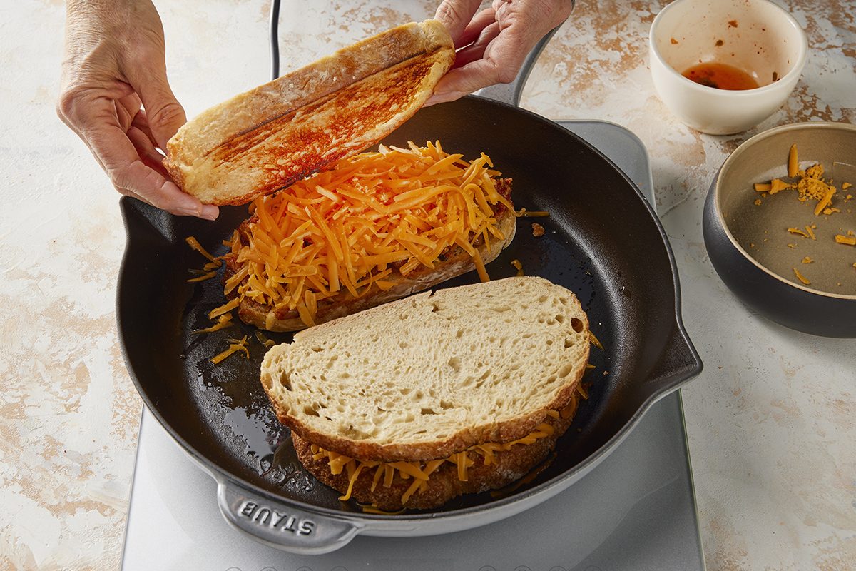 A person assembles a grilled cheese sandwich in a skillet, adding a slice of bread with sauce to a layer of shredded cheese and bread. Nearby are a bowl of sauce and a plate with leftover shredded cheese.
