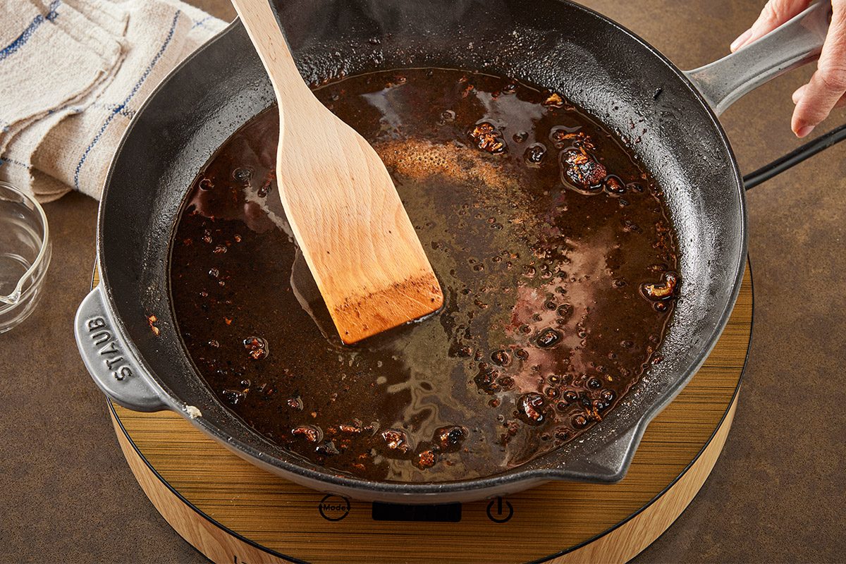 3/4 angle view shot of a hand stirs a dark sauce with a wooden spatula in a cast iron skillet on an electric burner and a kitchen towel sits close by while a small glass bowl rests nearby;