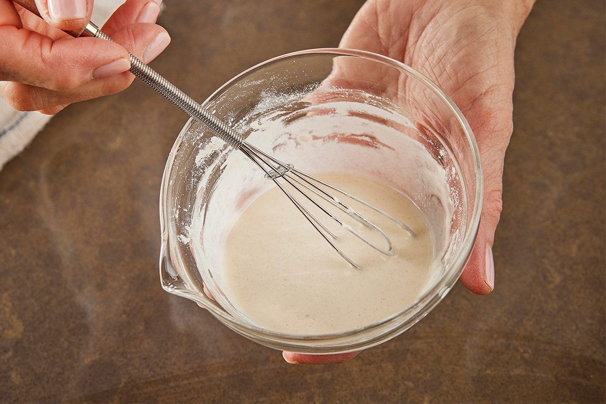 Close shot of a person holds a glass bowl of light-colored batter or sauce in one hand and mixes it with a small metal whisk using the other hand the motion appears gentle as the mixture swirls smoothly;