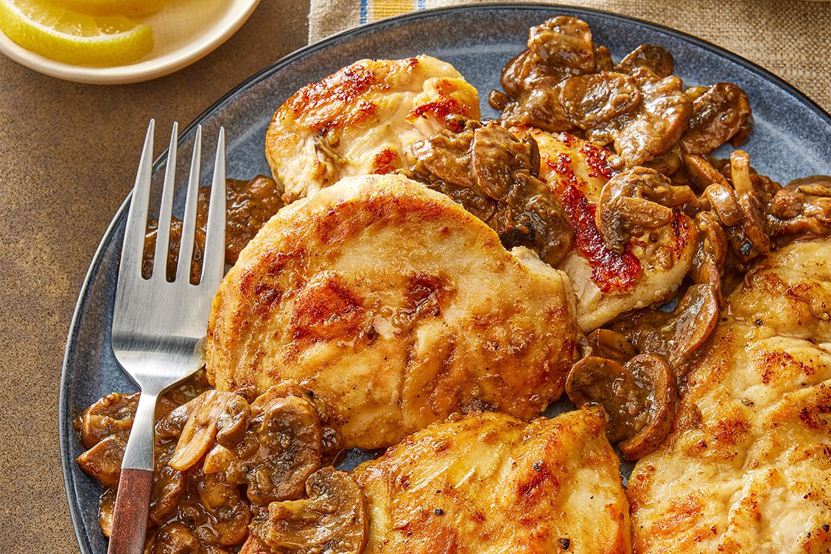 High angle view shot of Lemon Mushroom Chicken with three grilled chicken breasts topped with sautéed mushrooms; A fork is on the plate and a small dish of lemon wedges sits nearby on a beige and yellow striped cloth;