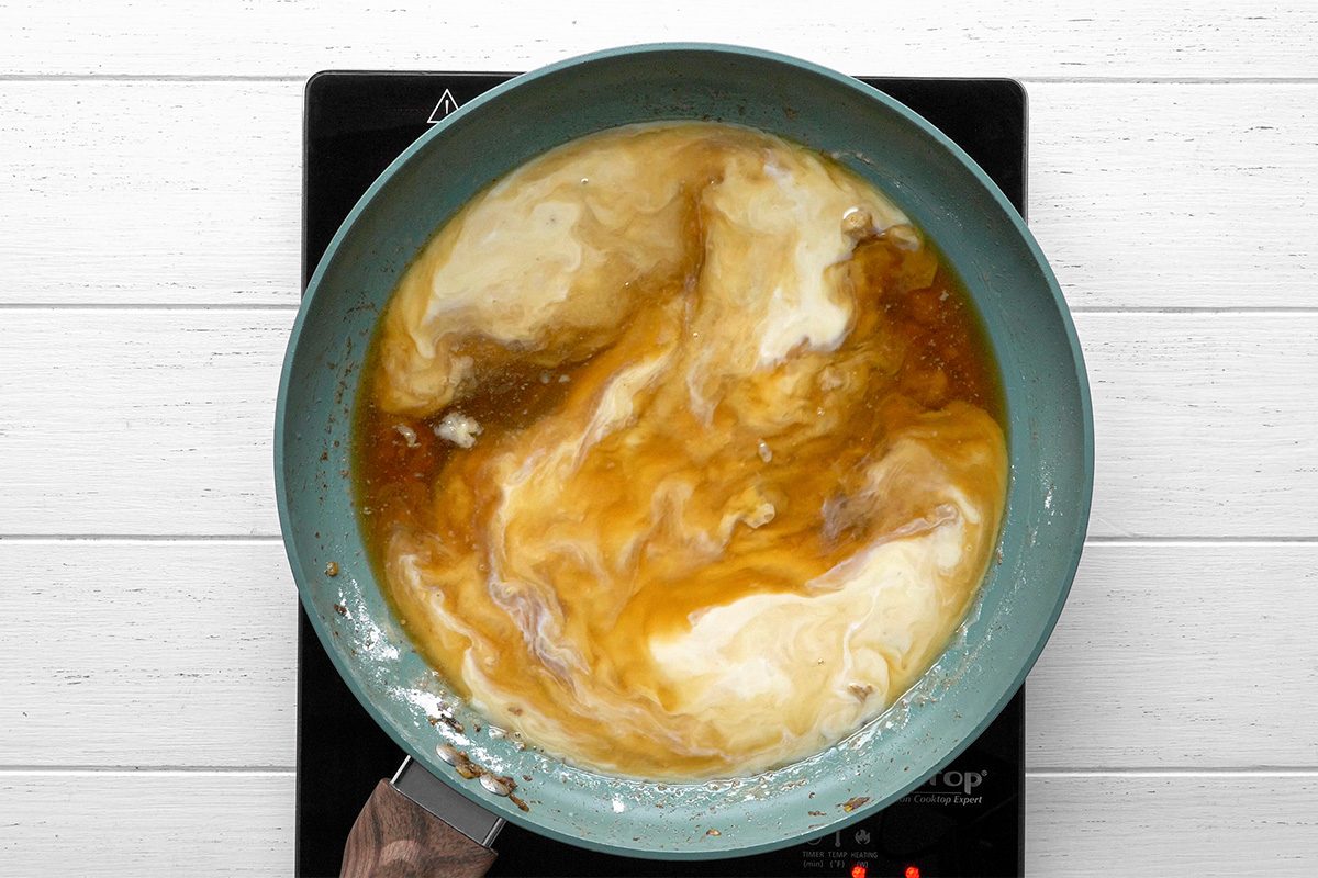 overhead shot of frying pan on an electric stove contains a swirling mixture of light and dark liquids, likely cream and broth, being stirred together over heat, The background is a white wooden surface