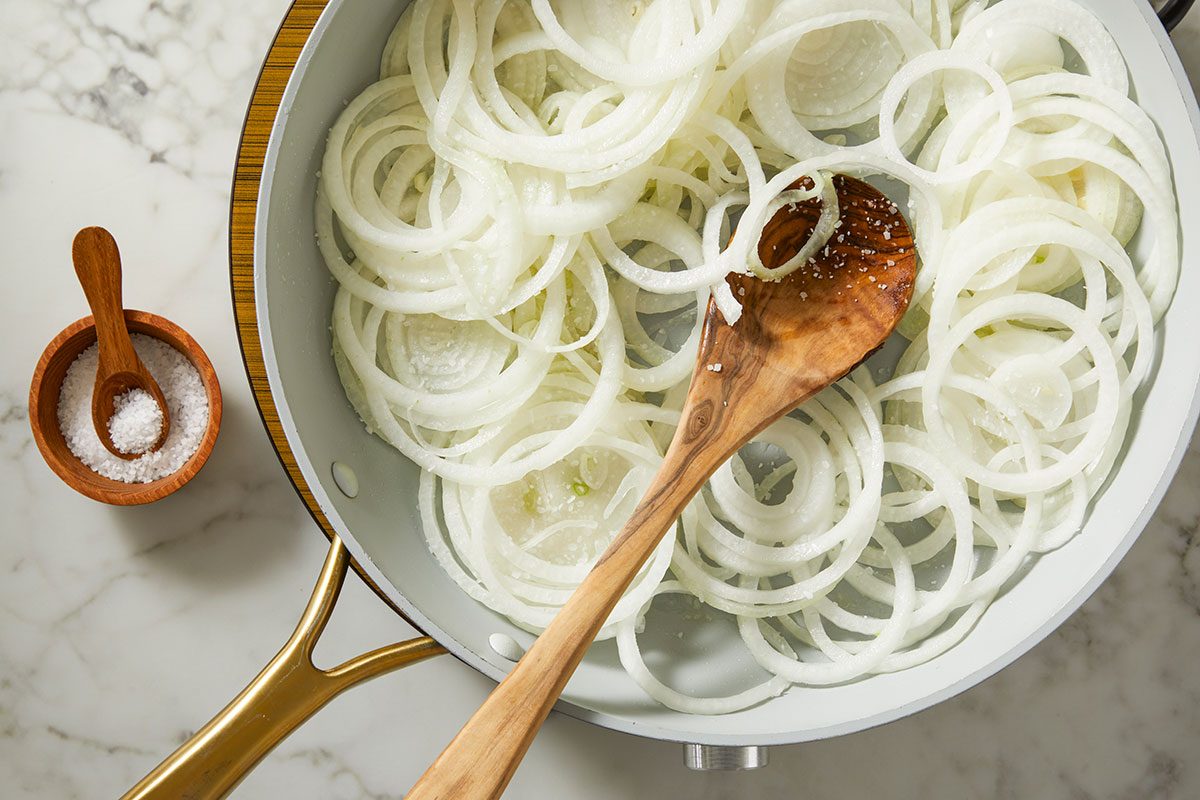 An overhead, horizontal step-by-step image showing thinly sliced sweet onions in a skillet with olive oil and butter, beginning the caramelization process for Million-Dollar Gnocchi.