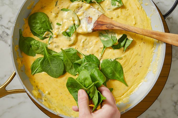 An overhead, horizontal step-by-step image showing fresh baby spinach being folded into the creamy gnocchi sauce for Million-Dollar Gnocchi.