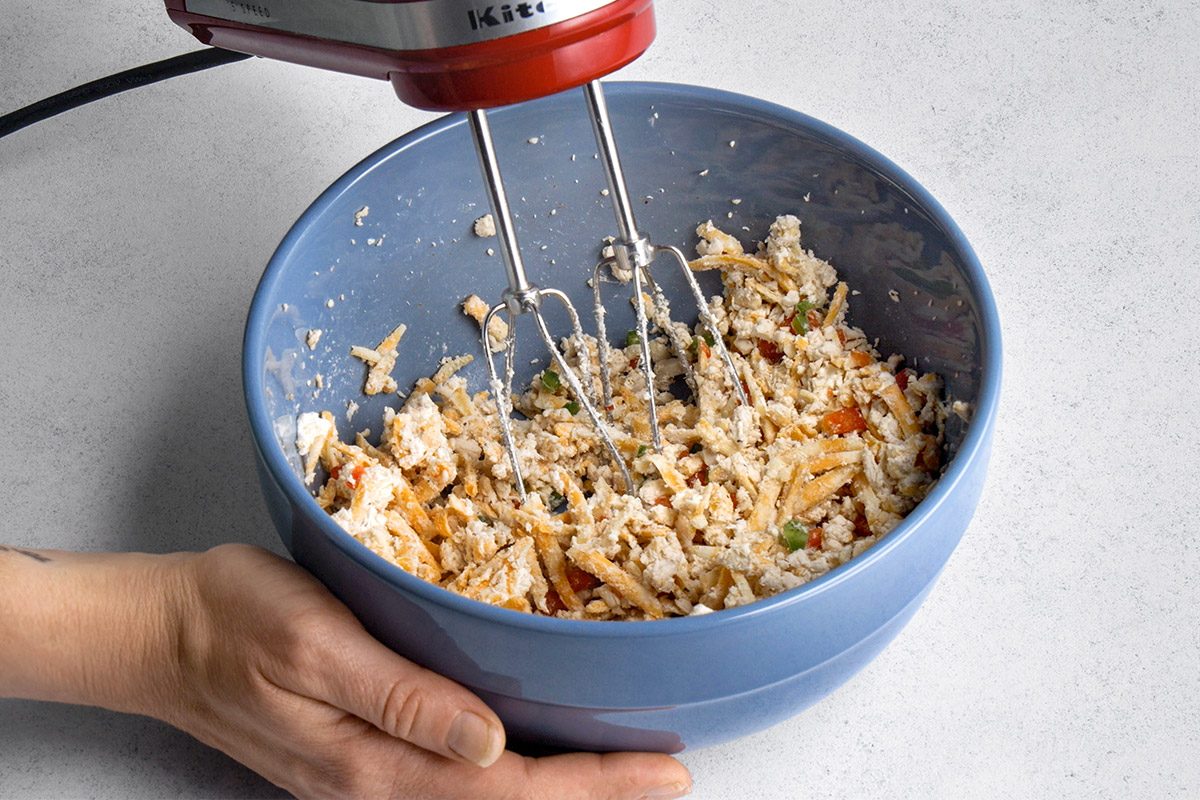 High angle shot of a person using a hand mixer to blend ingredients in a blue bowl; The mixture includes shredded chicken, cheese, and chopped vegetables;