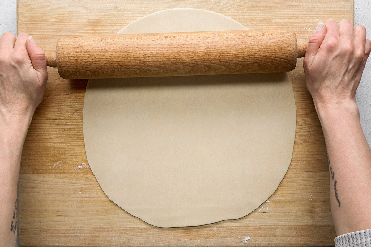 Overhead shot of hands rolling out dough with a wooden rolling pin on a wooden board, forming a round, flat sheet;