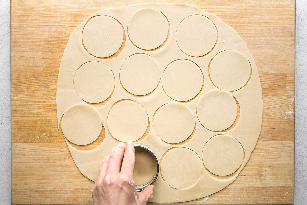 Overhead shot of hands rolling out dough with a wooden rolling pin on a wooden board, forming a round, flat sheet;