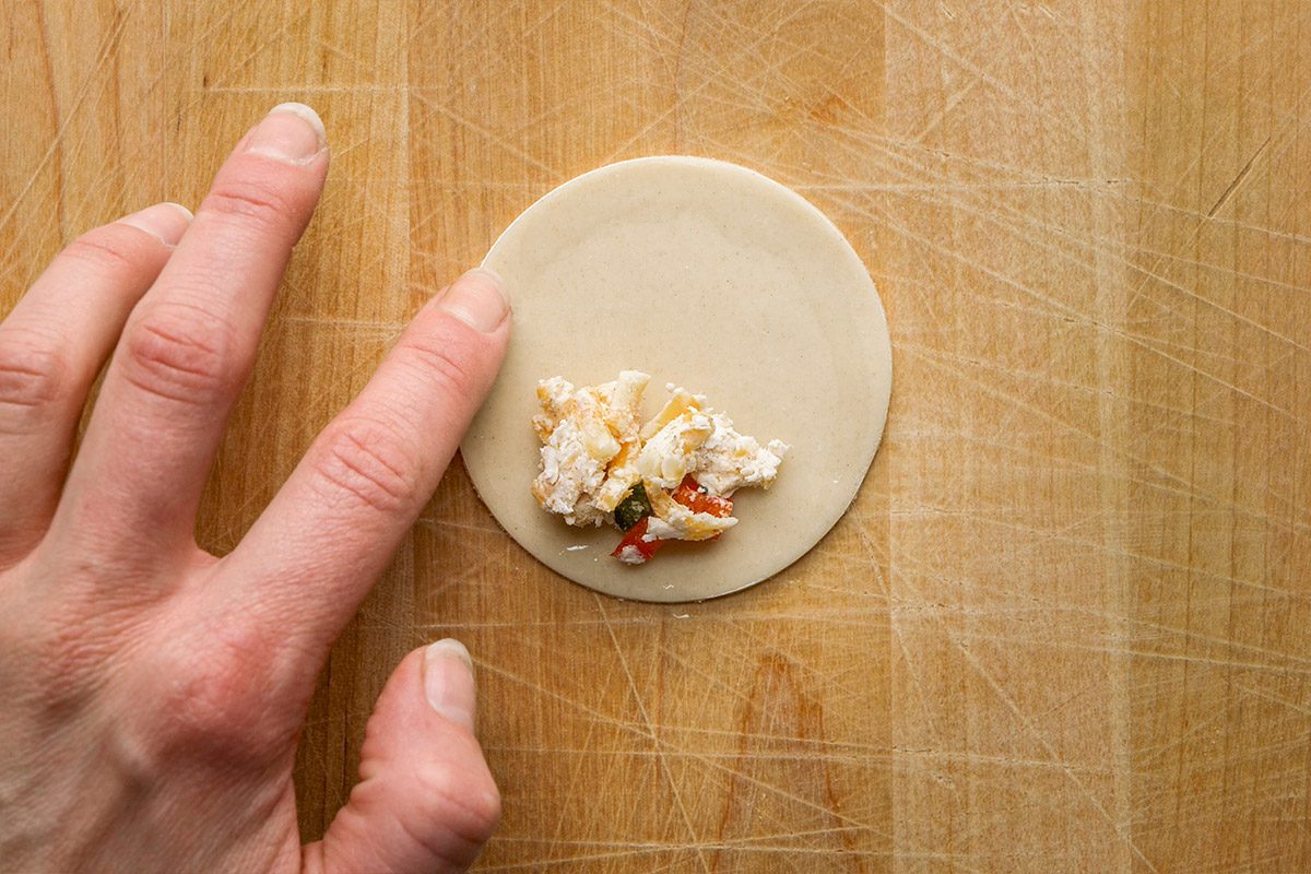 Overhead shot of a hand placing a small amount of vegetable filling onto a round piece of dough on a wooden surface, preparing to fold it for dumplings or pastries;