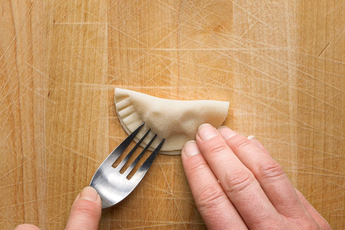 Overhead shot of a hand holding a half-moon-shaped dumpling on a wooden surface while pressing the edges with a fork to seal it;