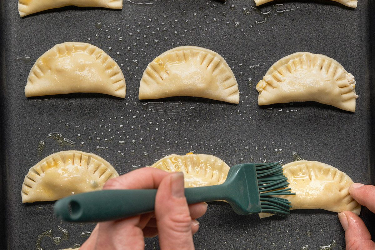 Overhead shot of a hand brushing egg wash onto semi-circular pastry turnovers arranged on a dark baking tray, ready for baking;