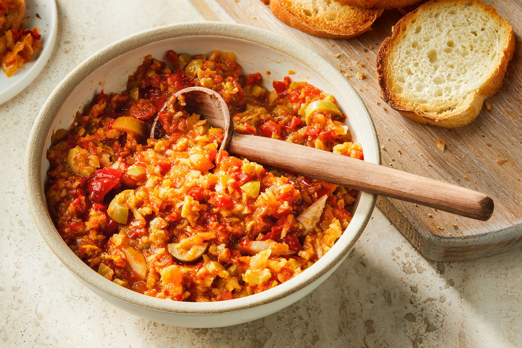 Olive Relish in a bowl with a serving spoon in it. Some bread placed on the side.