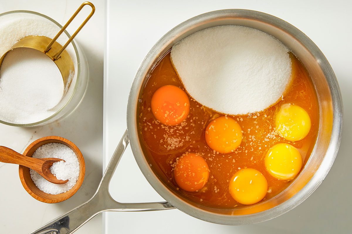 Overhead, horizontal step-by-step image of whole eggs, egg yolks, sugar, and thawed passion fruit juice concentrate combined in a saucepan as the base for the passion fruit tart filling.