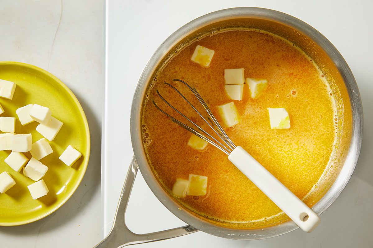 Overhead, horizontal step-by-step image showing cubed butter being added to a warm passion fruit custard mixture while whisking to create a smooth tart filling.