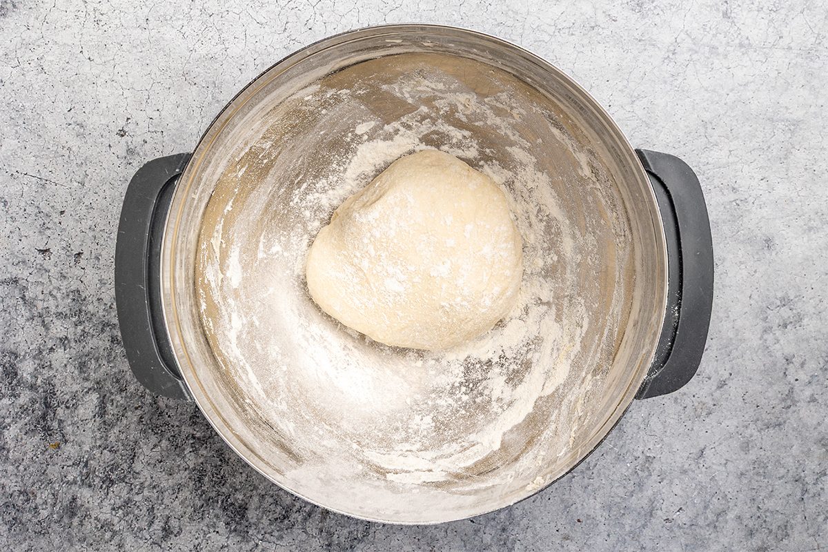 overhead shot of A ball of bread dough sits in the center of a floured metal mixing bowl, on a gray textured surface