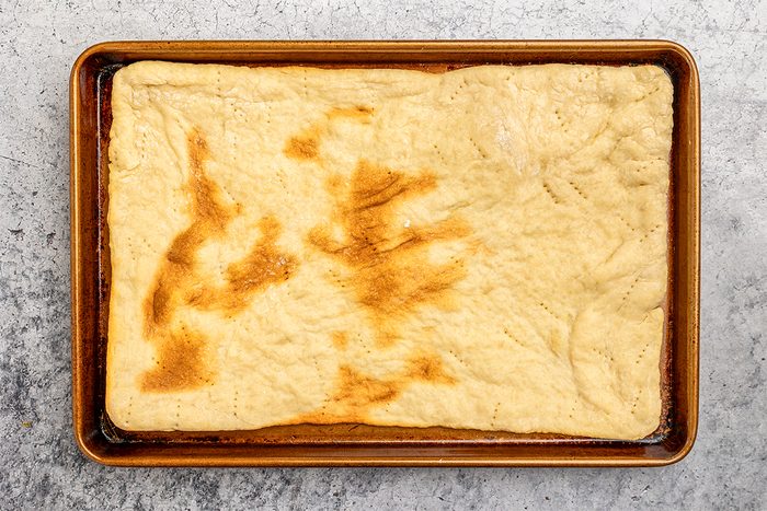 overhead shot of A rectangular sheet of baked dough with uneven brown spots sits on a rimmed metal baking tray, placed on a gray textured surface