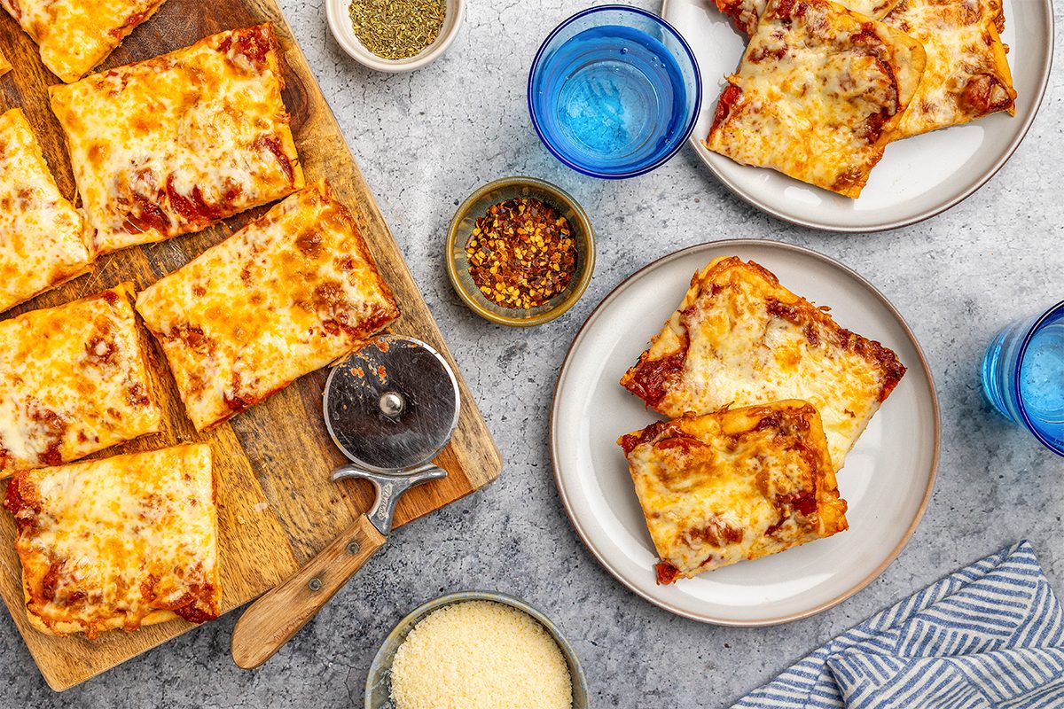 overhead shot of Rectangular slices of cheesy pizza on a wooden board and plates, with a pizza cutter, grated cheese, chili flakes, dried herbs, and blue glasses of water on a gray surface; A striped napkin is at the lower right
