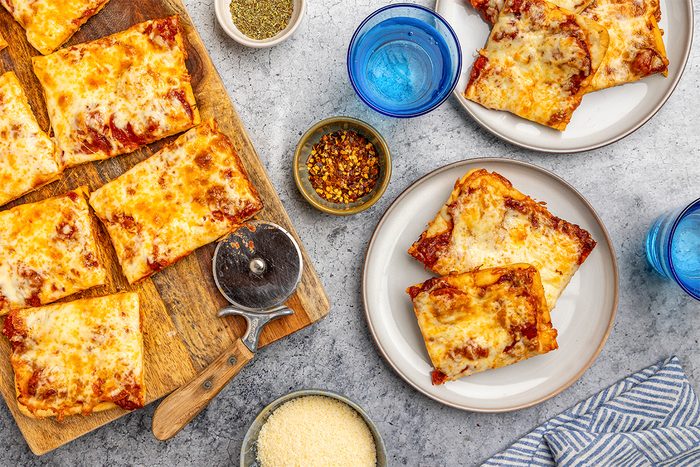 overhead shot of Rectangular slices of cheesy pizza on a wooden board and plates, with a pizza cutter, grated cheese, chili flakes, dried herbs, and blue glasses of water on a gray surface; A striped napkin is at the lower right