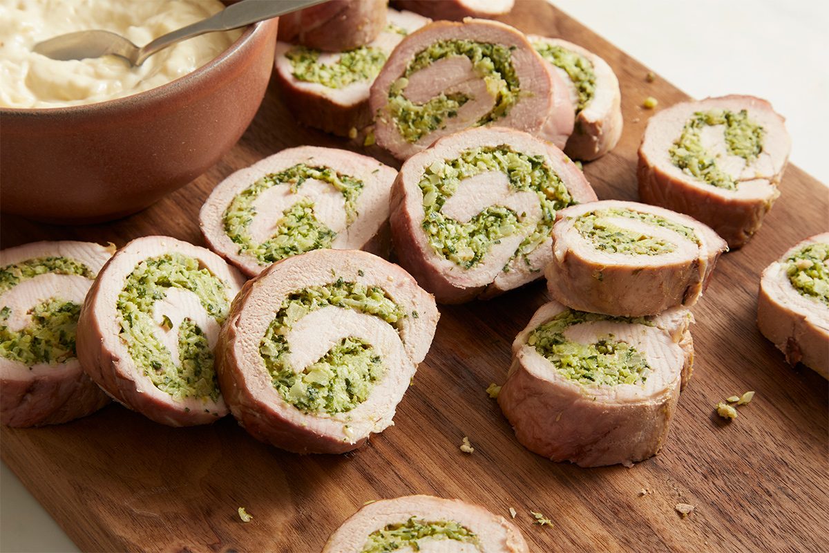 Close up shot of a wooden board displaying seared Pork Pinwheels slices, with a bowl of creamy sauce visible in the background.