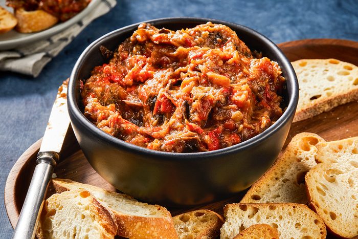 A black bowl filled with chunky vegetable and tomato dip is surrounded by slices of toasted bread on a wooden tray, with a knife resting nearby.