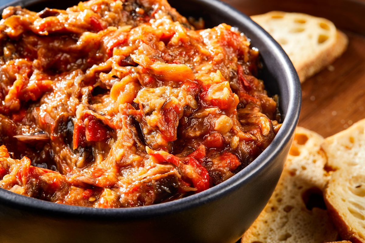 A close-up of a bowl filled with chunky, reddish-orange eggplant dip, surrounded by slices of crusty bread on a wooden surface.