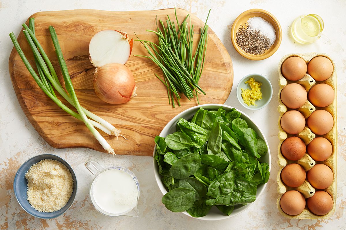 A wooden board with green onions, a yellow onion, and chives sits beside bowls of spinach, grated cheese, cream, lemon zest, black pepper, oil, and a carton of eggs on a light countertop.
