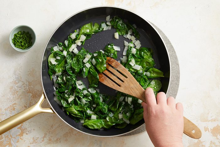 A hand stirs chopped spinach and diced onions in a frying pan with a wooden spatula. A small bowl of chopped herbs sits nearby on a light-colored surface.