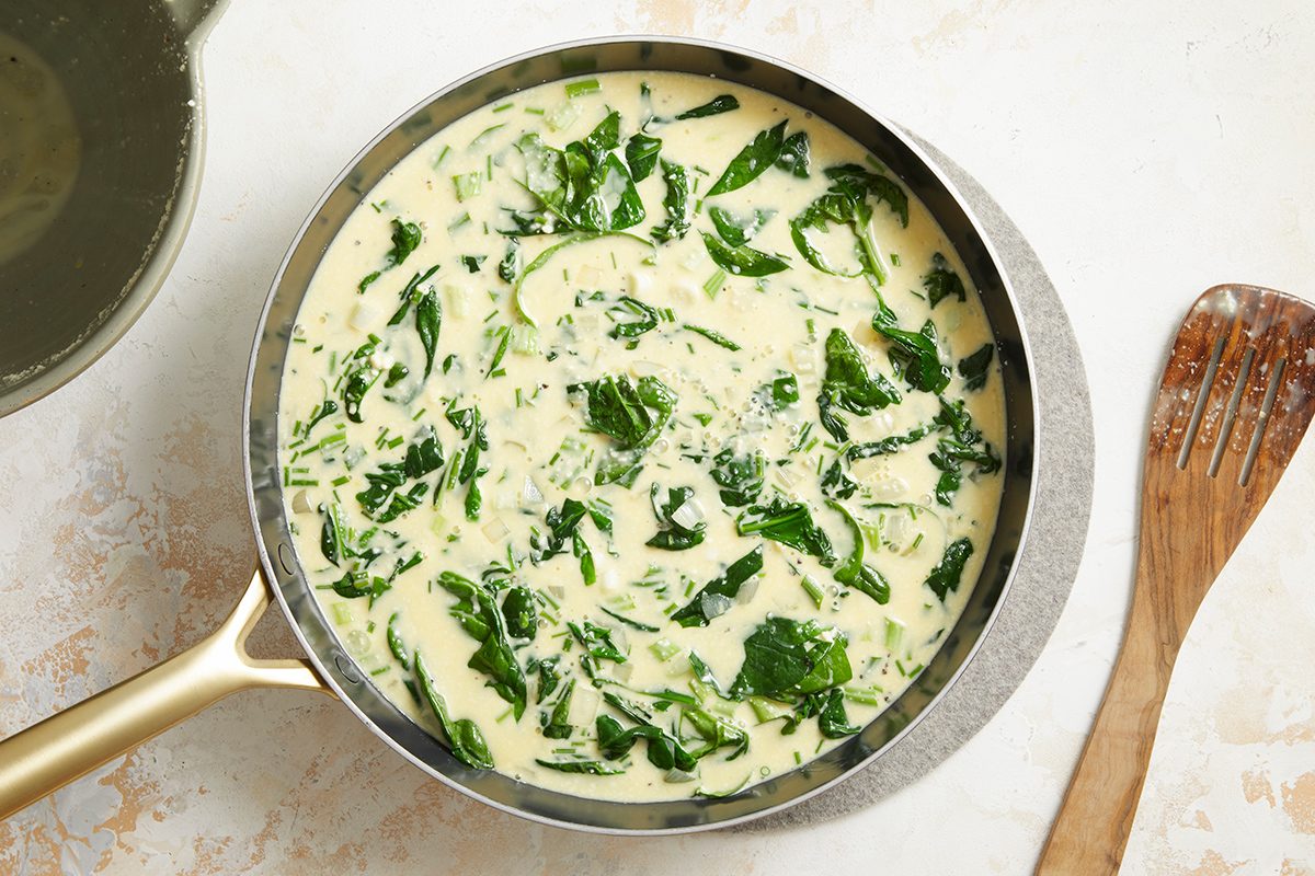 A pan filled with creamy spinach sauce sits on a light surface beside a wooden spatula. The sauce is pale yellow and mixed with vibrant green spinach leaves.