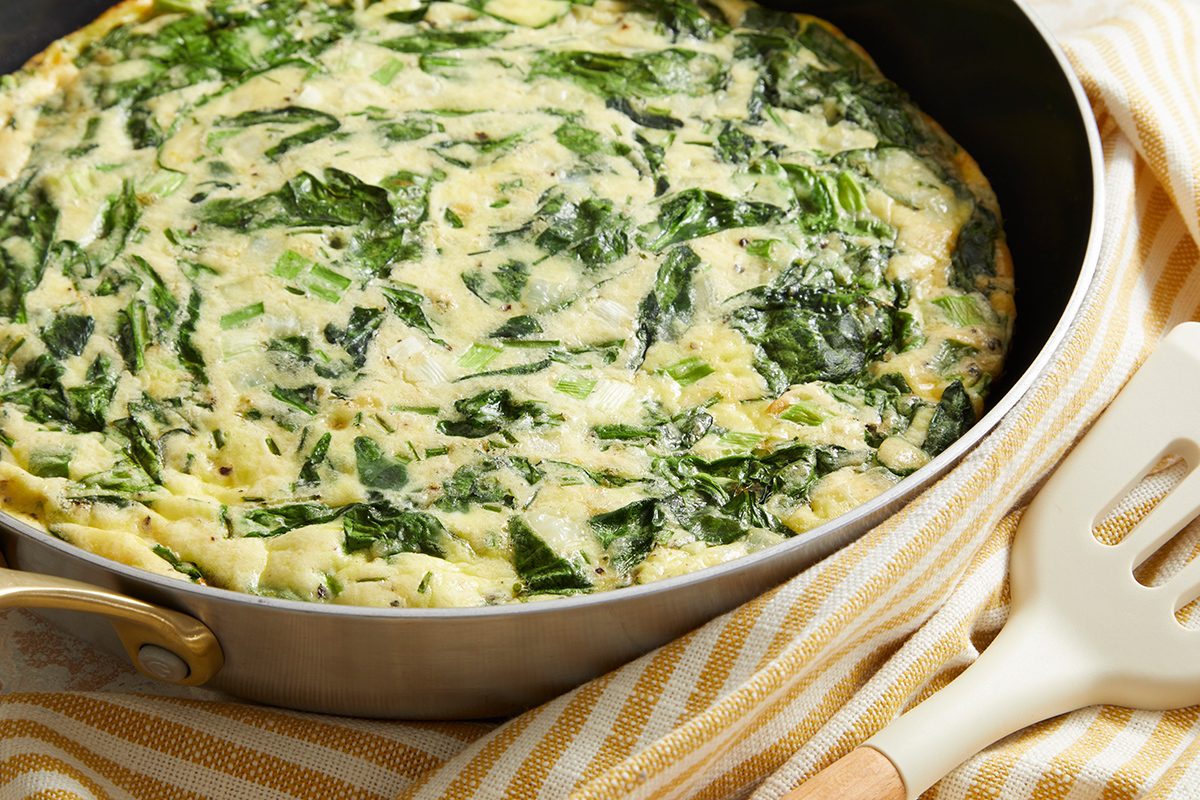 A close-up of a spinach and cheese frittata in a pan, resting on a beige striped cloth with a white spatula next to it. The frittata appears golden and filled with leafy spinach.