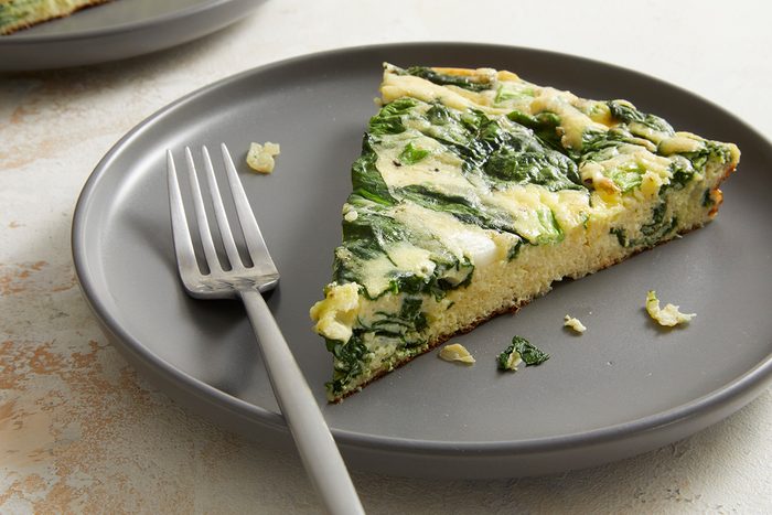 A slice of spinach and cheese frittata sits on a gray plate with a silver fork, with a few crumbs around the slice.