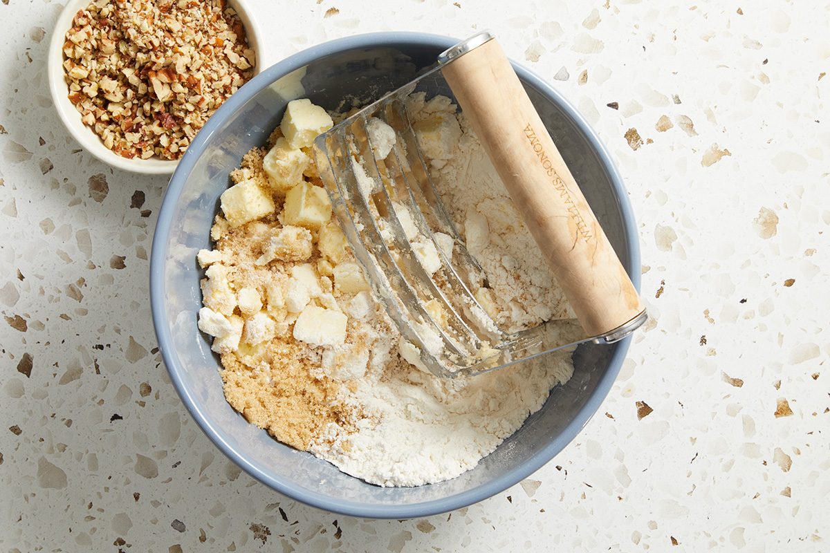 A bowl with flour, cubed butter, and brown sugar being mixed with a pastry cutter, with a small bowl of chopped nuts beside it on a speckled countertop.