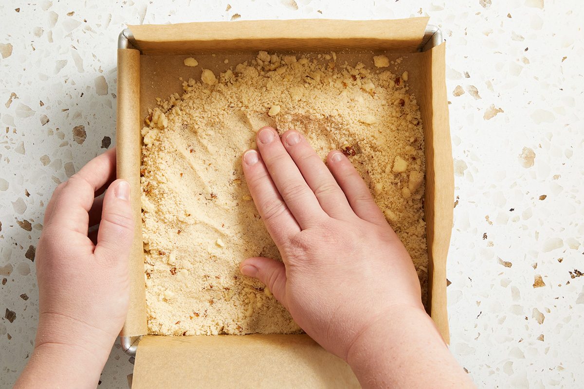 A person presses a crumbly crust mixture into the bottom of a square baking pan lined with parchment paper.