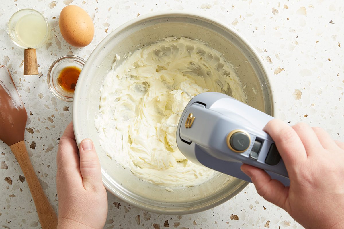 A person uses a hand mixer to blend cream cheese in a bowl, with an egg, vanilla, lemon juice, and a spatula on a speckled countertop nearby.