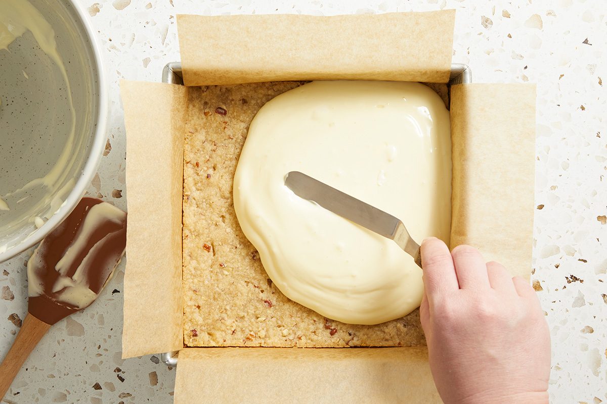A hand uses a spatula to spread creamy white frosting over a square baked crust in a parchment-lined pan, with a mixing bowl and spatula nearby on a speckled countertop.