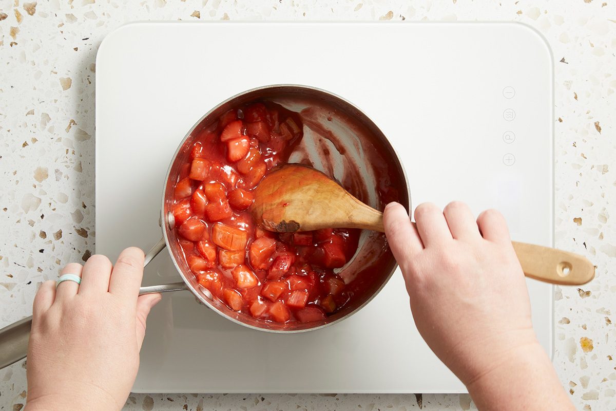 A person stirs chopped strawberries in a saucepan with a wooden spoon, cooking the mixture on a white stovetop.