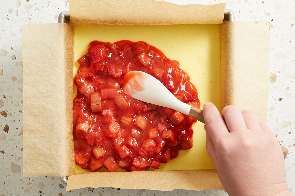 A hand uses a white spatula to spread a chunky red fruit filling over a yellow dough base in a parchment-lined square baking pan.