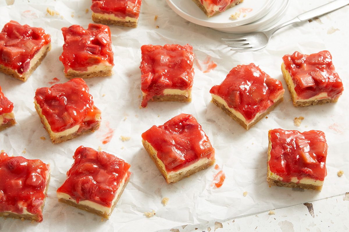 Several square strawberry-topped dessert bars are arranged on a sheet of parchment paper, with a plate and forks in the background. The bars have a crumbly crust and glossy strawberry topping.