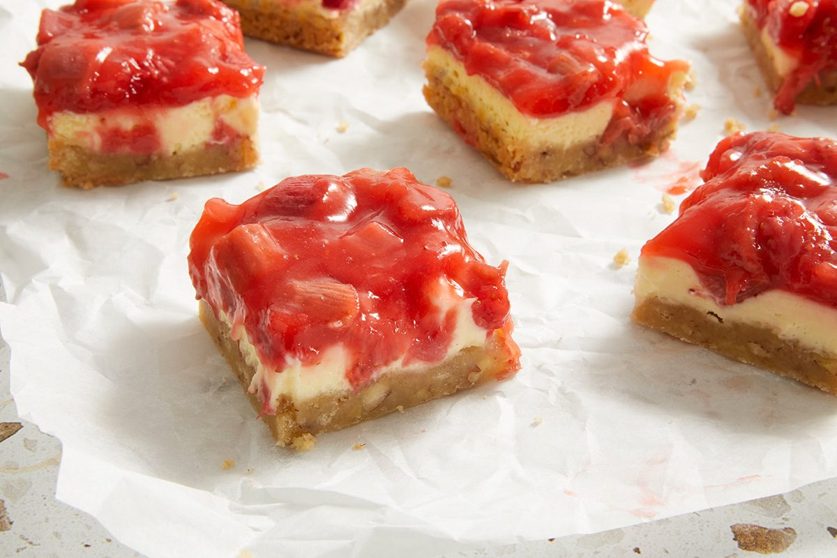 Several strawberry cheesecake bars with a golden brown crust, creamy cheesecake layer, and glossy strawberry topping are arranged on a sheet of white parchment paper.