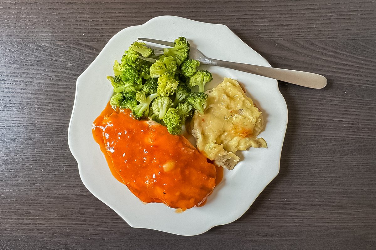 A white plate with broccoli florets, cheesy scalloped potatoes, and a piece of meatloaf topped with red sauce, placed on a dark wooden table with a fork.