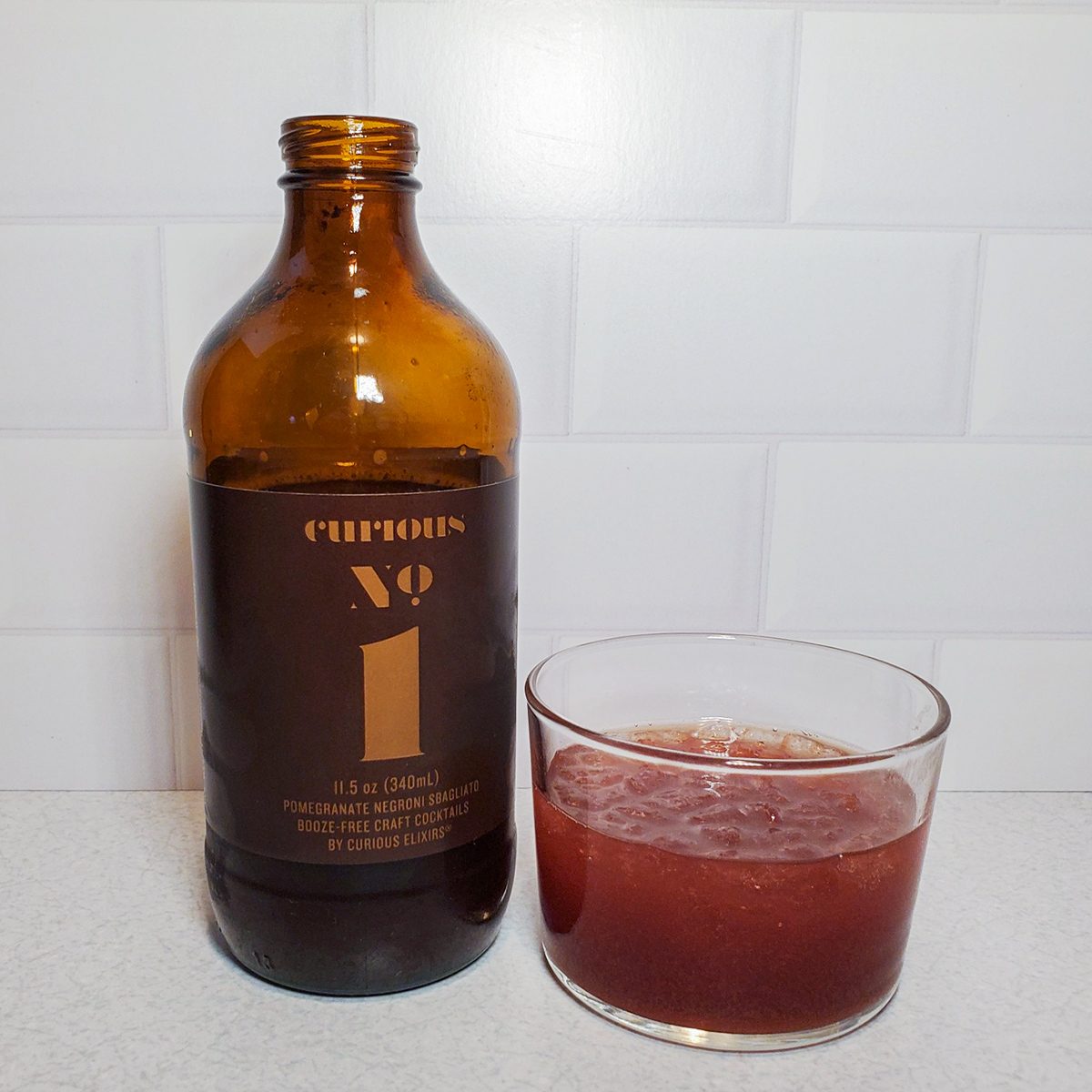 A brown glass bottle labeled "Curious No. 1" stands next to a small glass filled with a reddish-brown beverage on a light countertop, with a white subway tile background.