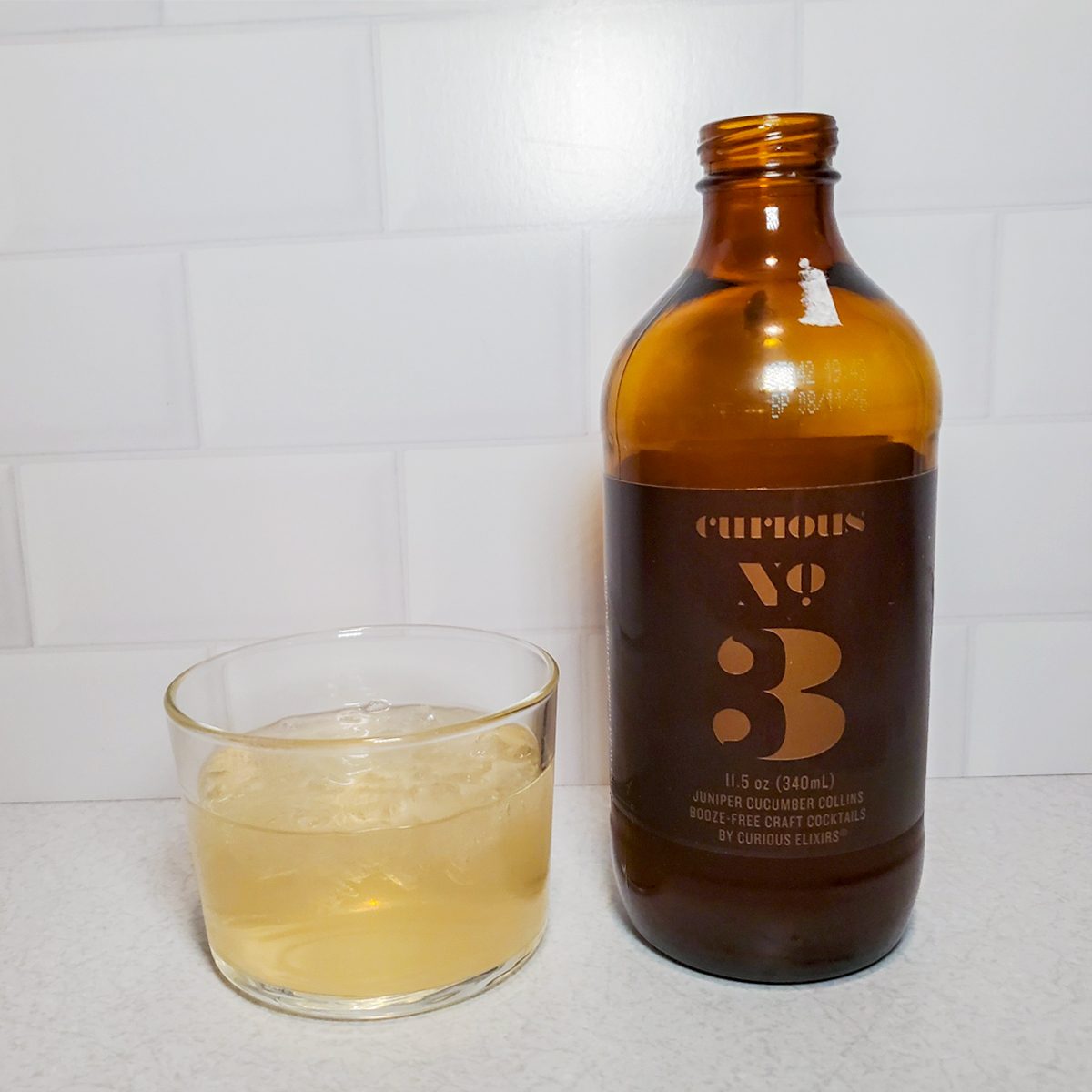 A glass filled with a light-colored drink and ice sits next to a brown bottle labeled “Curious No. 3” on a white countertop with a white tiled background.