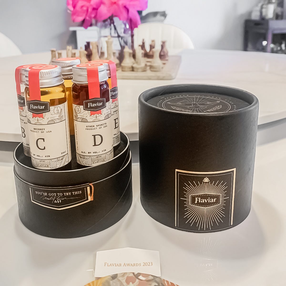 A Flaviar tasting box with three labeled vials of spirits is displayed on a table. The open black cylindrical Flaviar container is beside the vials, with a blurred background of a chessboard and flowers.