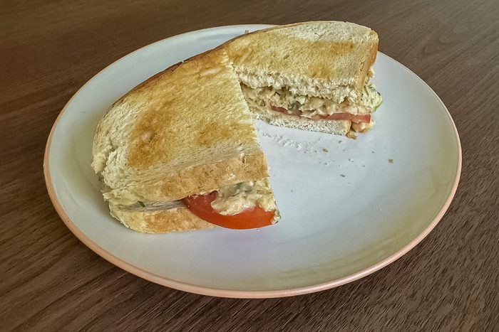 A toasted sandwich cut in half on a white plate with a pink rim, showing a filling of sliced tomato and what appears to be chicken or egg salad. The plate rests on a wooden surface.