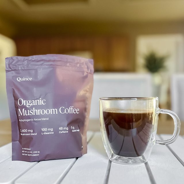 A clear glass mug of black coffee sits on a white table next to a purple bag labeled "Quince Organic Mushroom Coffee" in a bright indoor setting with blurred background.