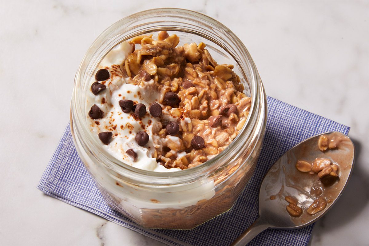 3/4 shot of a single jar of Tiramisu Overnight Oats topped with chocolate chips and cocoa powder, resting on a folded linen napkin with a spoon beside it.